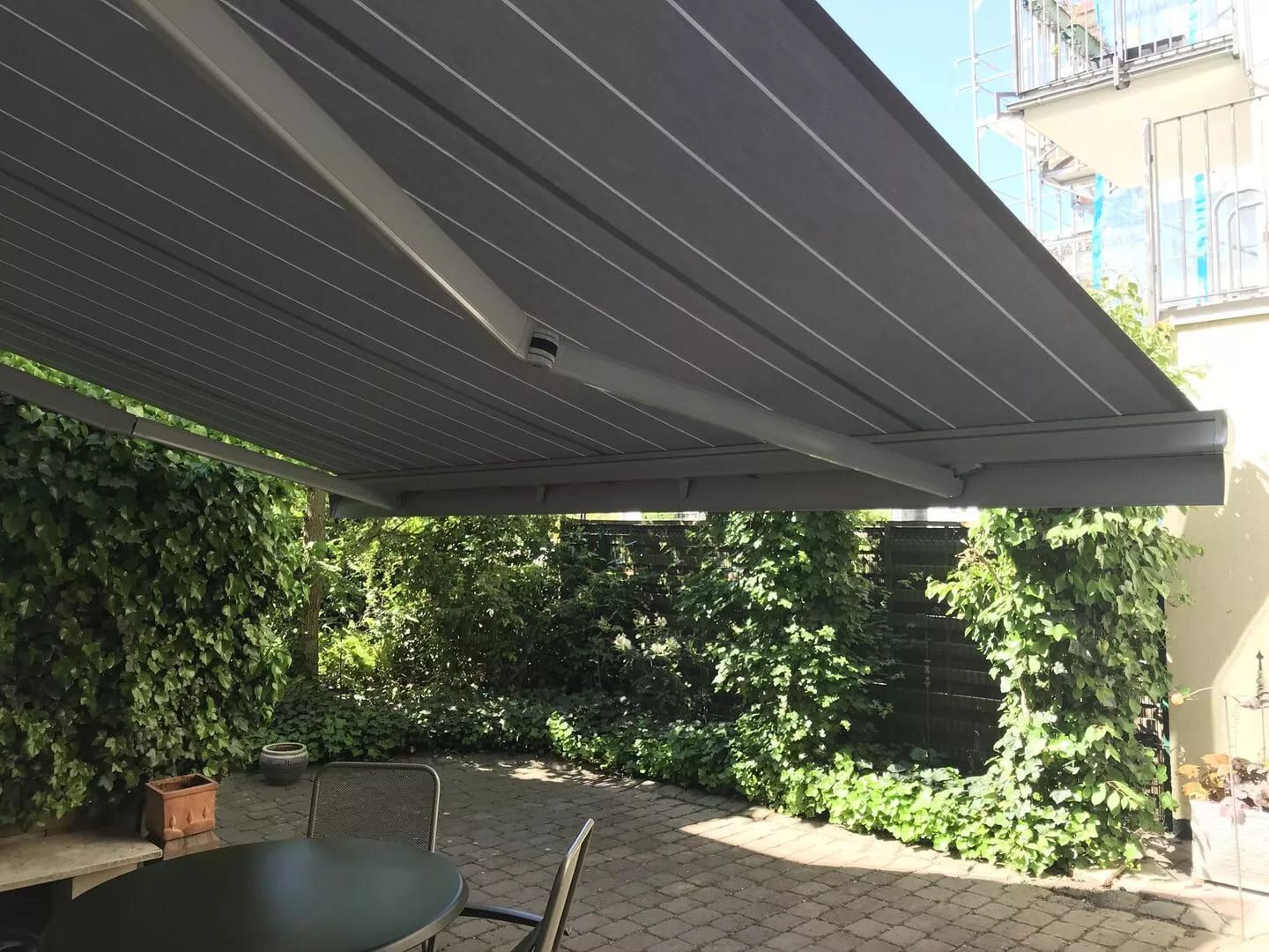 Eine versenkbare graue Markise spendet Schatten über einer Terrasse mit grünem Efeu und einem kleinen Tisch, umgeben von üppigem Grün.