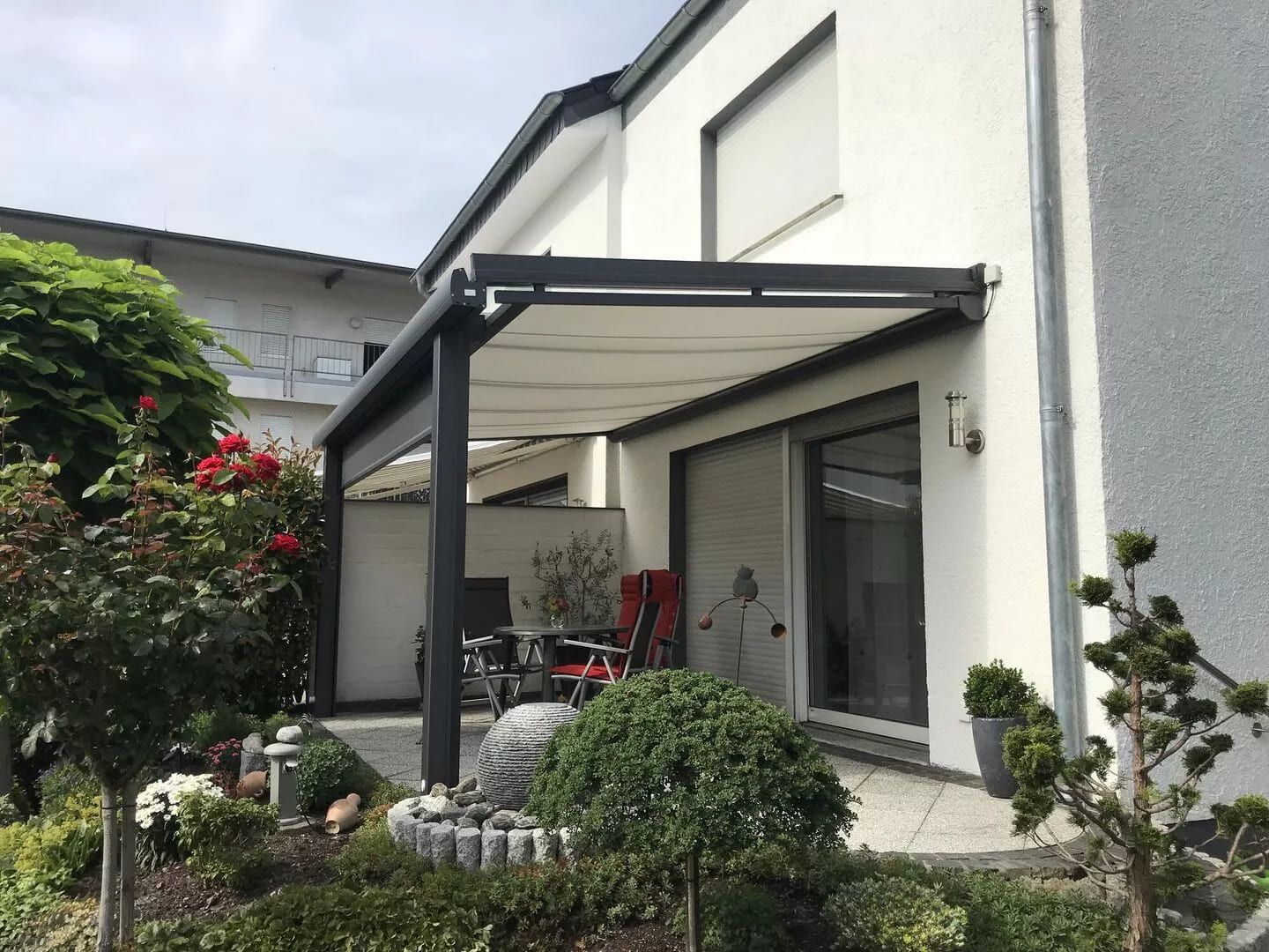 Eine moderne Terrasse mit weißer Markise, umgeben von üppigem Grün, Blumen und einem Essplatz im Freien zum Entspannen.