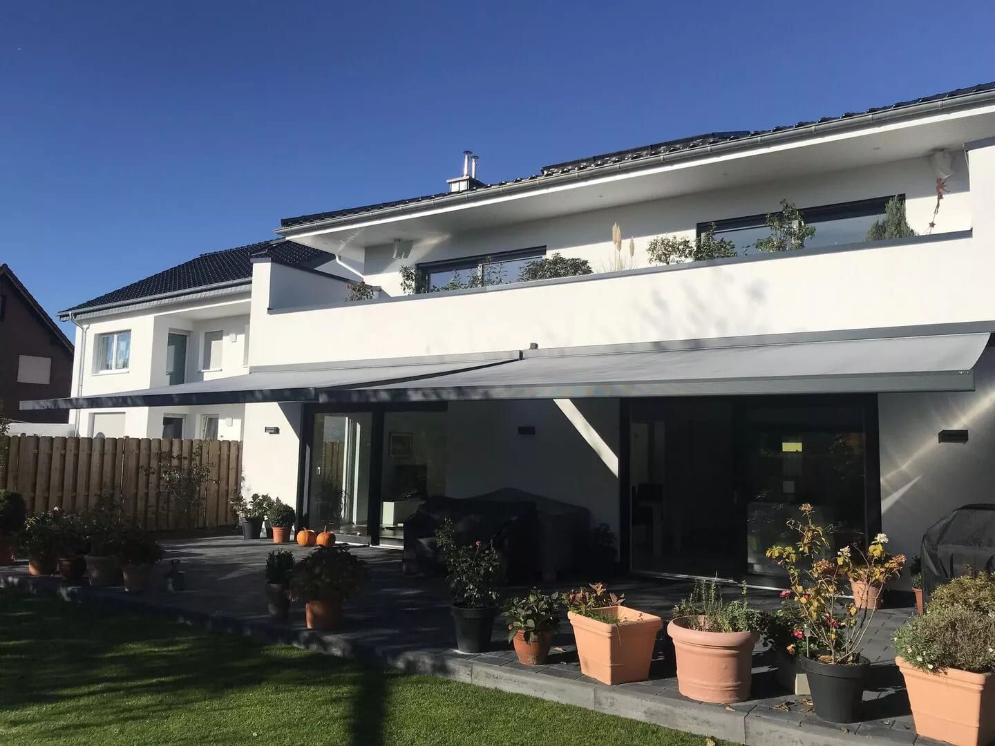 Modernes, zweistöckiges Haus mit großer Terrasse, Markise und verschiedenen Topfpflanzen unter einem strahlend blauen Himmel.