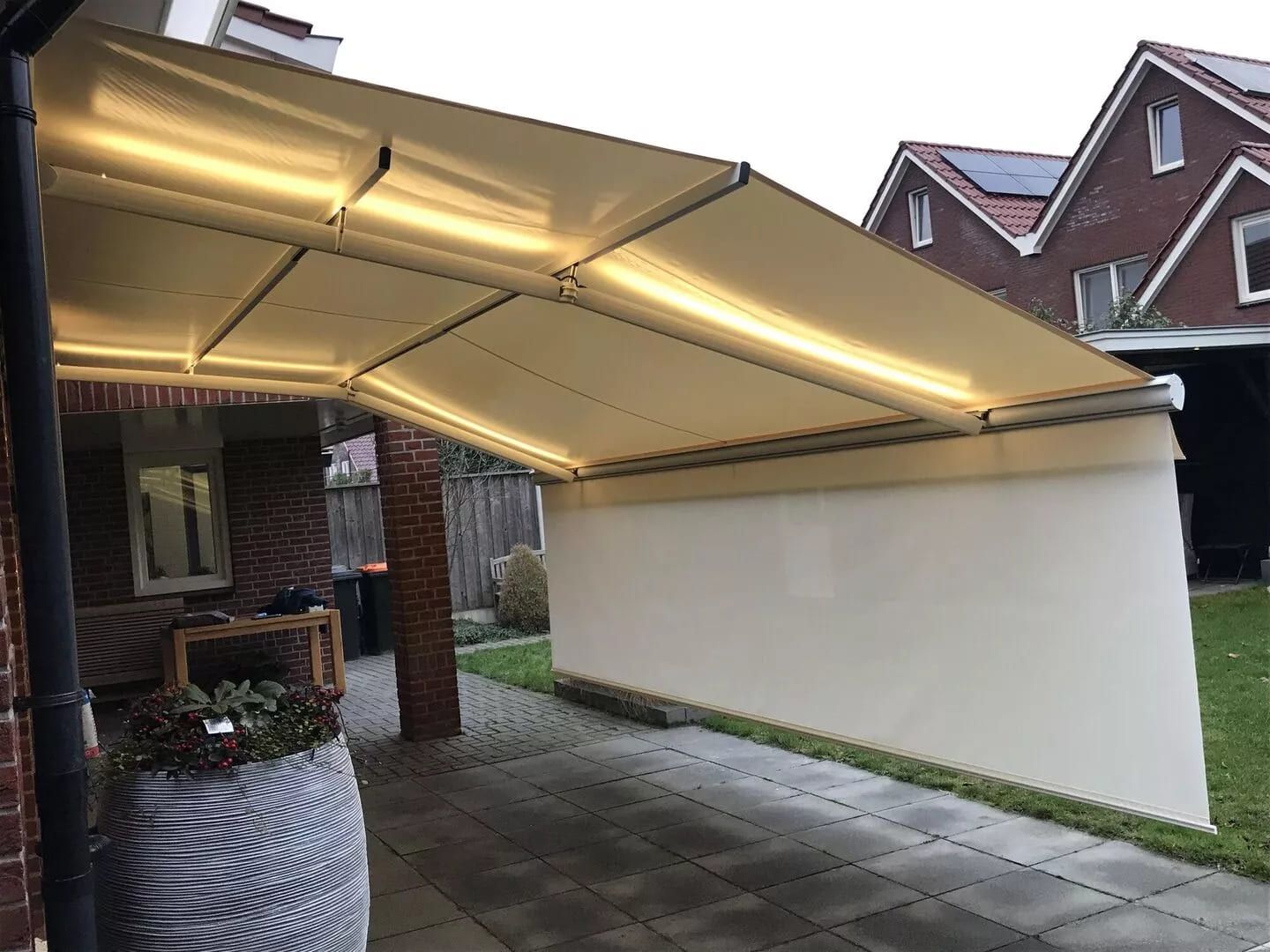 Eine moderne einziehbare Markise mit LED-Beleuchtung erstreckt sich über eine Terrasse und spendet Schatten und Atmosphäre in einem Hinterhof.