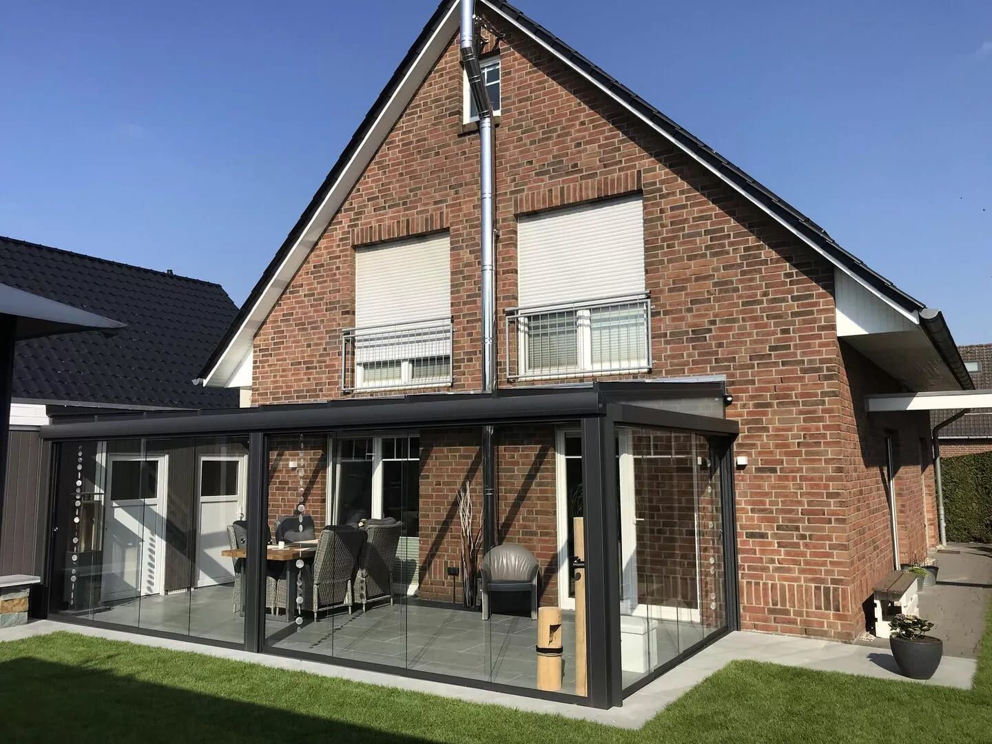 Ein modernes Backsteinhaus mit einem verglasten Innenhof mit Sitzgelegenheiten im Freien und einem grünen Rasen unter blauem Himmel.