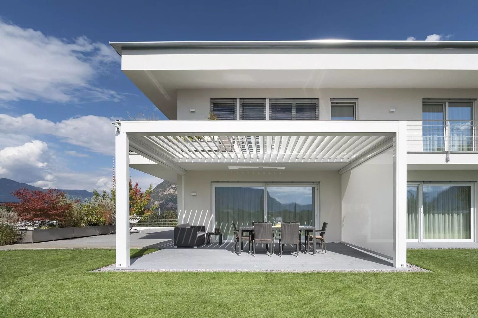 Ein modernes Haus mit einem geräumigen Innenhof mit einem gemütlichen Essbereich unter einer stilvollen Pergola, umgeben von üppigem grünen Rasen.