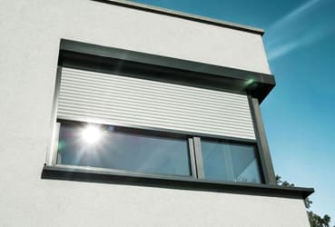 Ein modernes Fenster mit geschlossenem weißen Rollladen, das sich vor einem strahlend blauen Himmel im Sonnenlicht sonnt.