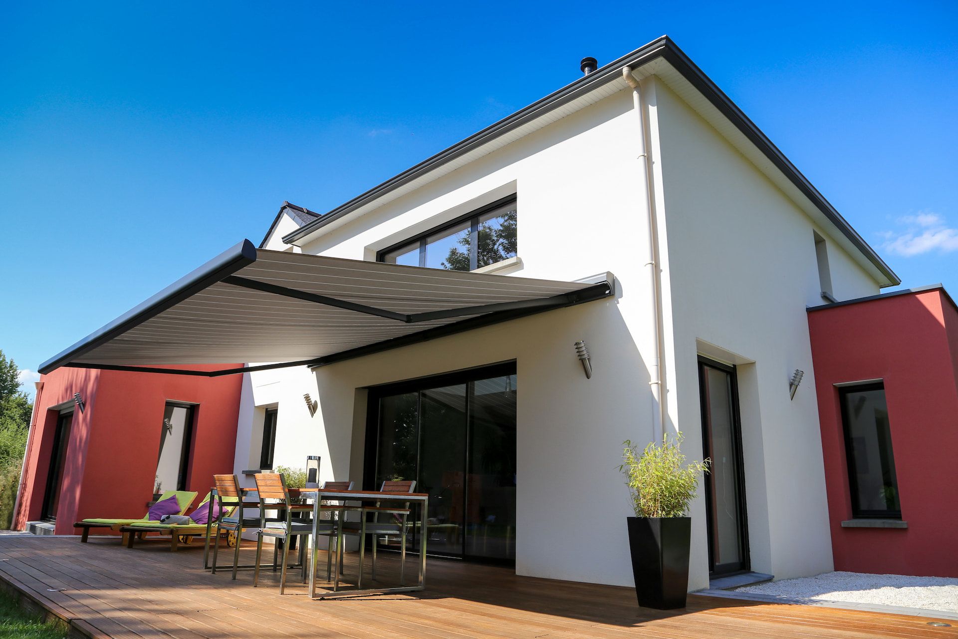 Modernes zweistöckiges Haus mit einer roten Akzentwand, großen Glastüren, einer Markise und einem Essbereich im Freien auf einer Holzterrasse.