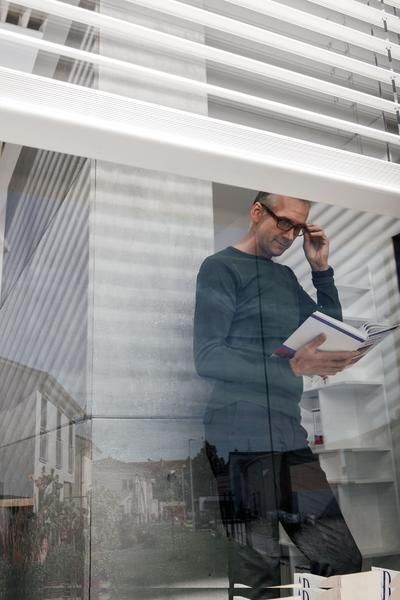 Eine Person steht in einem Haus und liest ein Buch, während sie nachdenklich aus einem großen Glasfenster schaut, durch das das Sonnenlicht Schatten wirft.