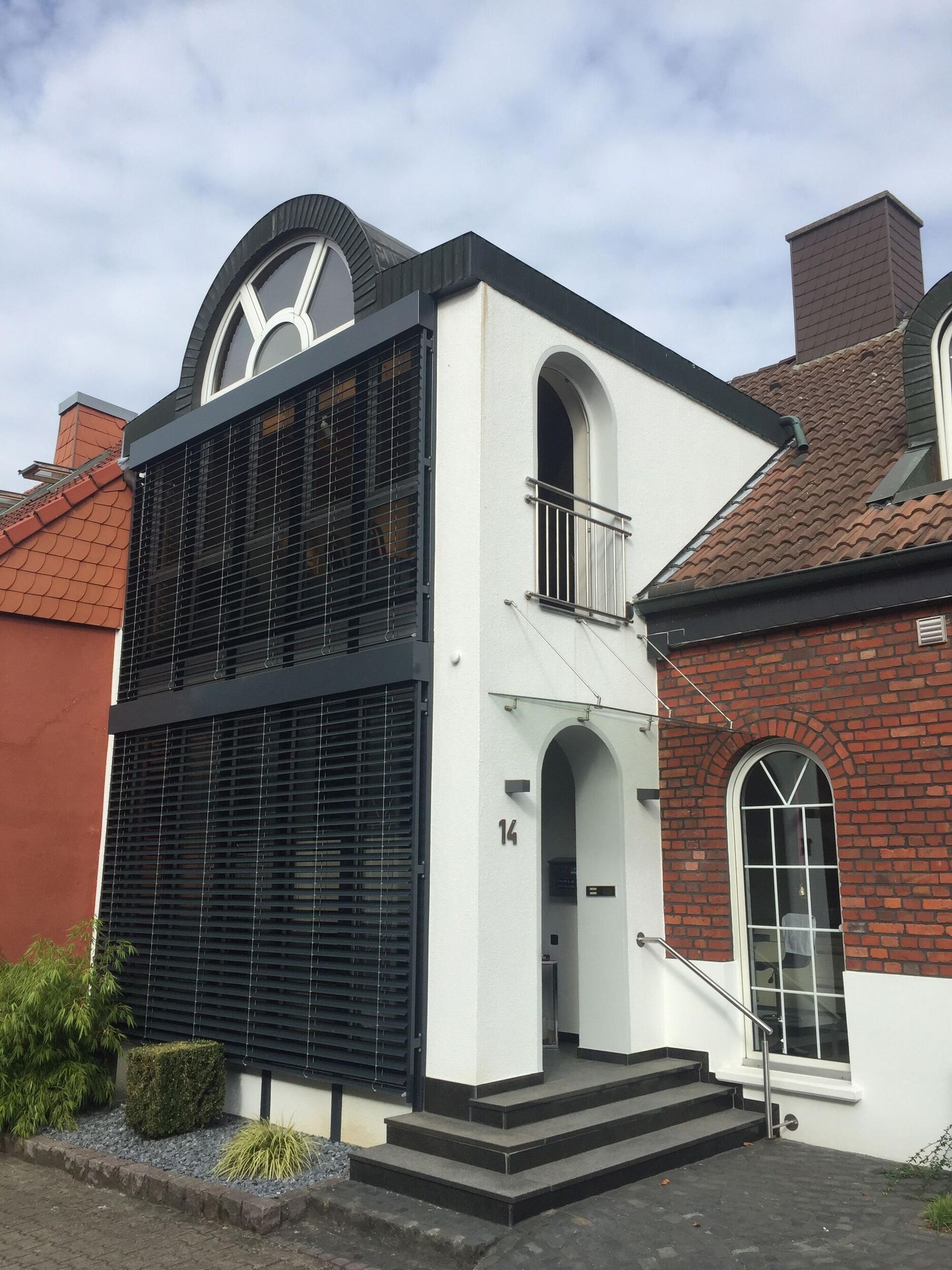 Modernes zweistöckiges Gebäude mit geschwungenen Fenstern und vertikalen Jalousien, das an ein klassisches Backsteinhaus angrenzt und über einen einladenden Eingang verfügt.