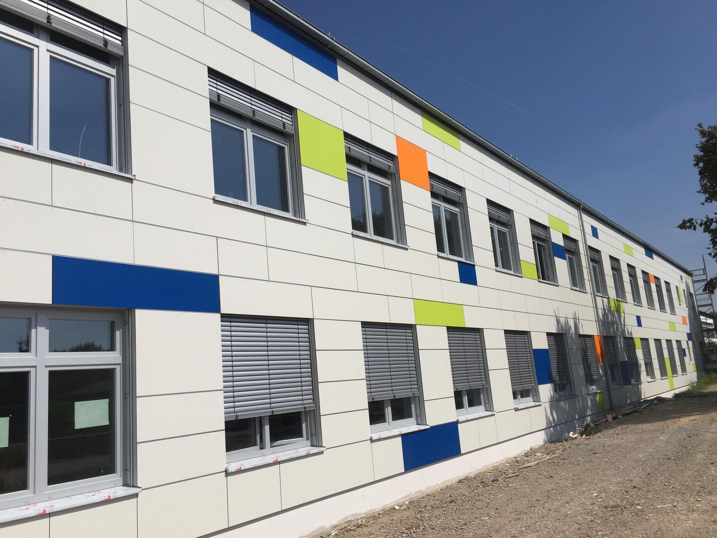 Eine moderne Gebäudefassade mit weißen Paneelen, die durch bunte blaue, grüne und orangefarbene Quadrate unter einem klaren blauen Himmel akzentuiert werden.