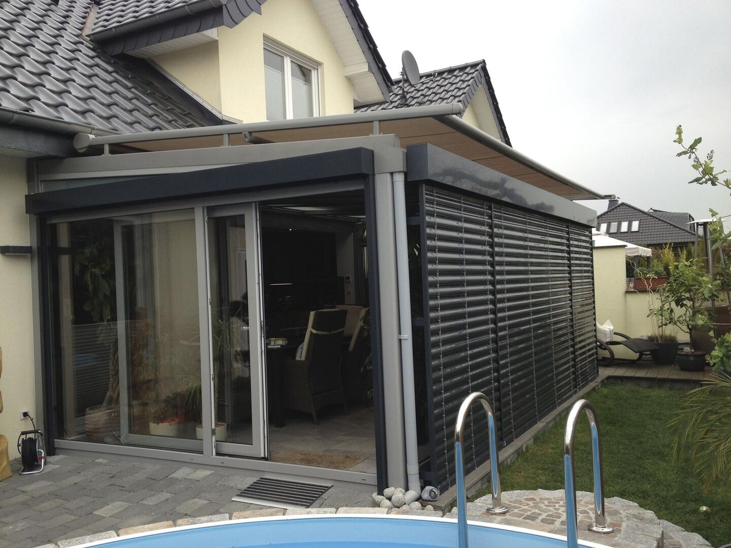 Moderner Hausanbau mit Glastüren und horizontalen Lamellen, mit Blick auf einen Garten mit Pool, unter bewölktem Himmel.