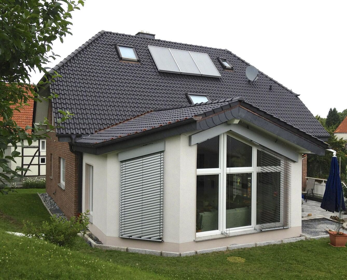 Modernes zweistöckiges Haus mit Schrägdach, großen Fenstern und einem begrünten Garten, mit Jalousien und Sonnenkollektoren auf dem Dach.