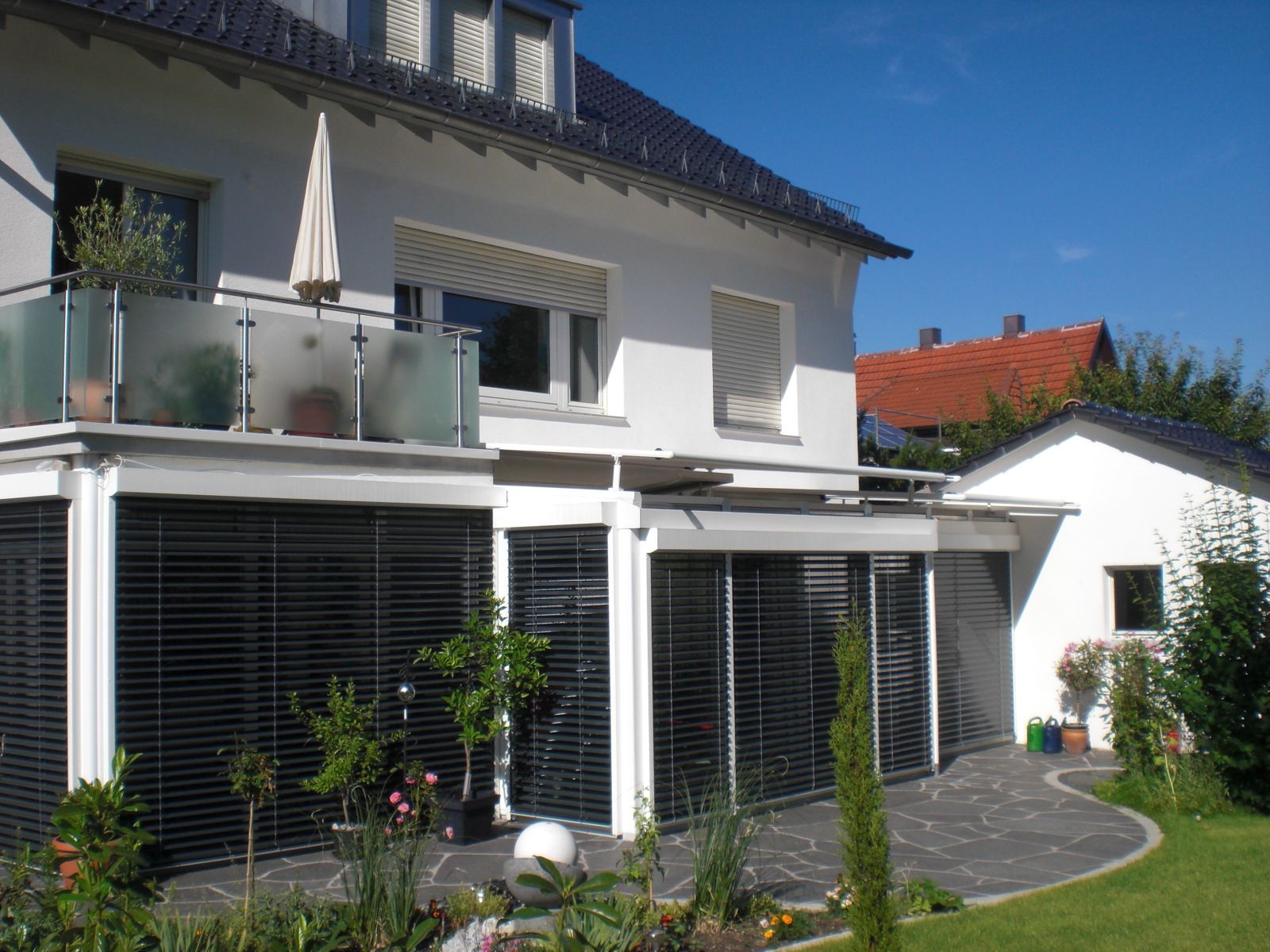 Modernes zweistöckiges Haus mit Balkon, großen Fenstern, schwarzen Fensterläden und einem gepflegten Garten unter strahlend blauem Himmel.