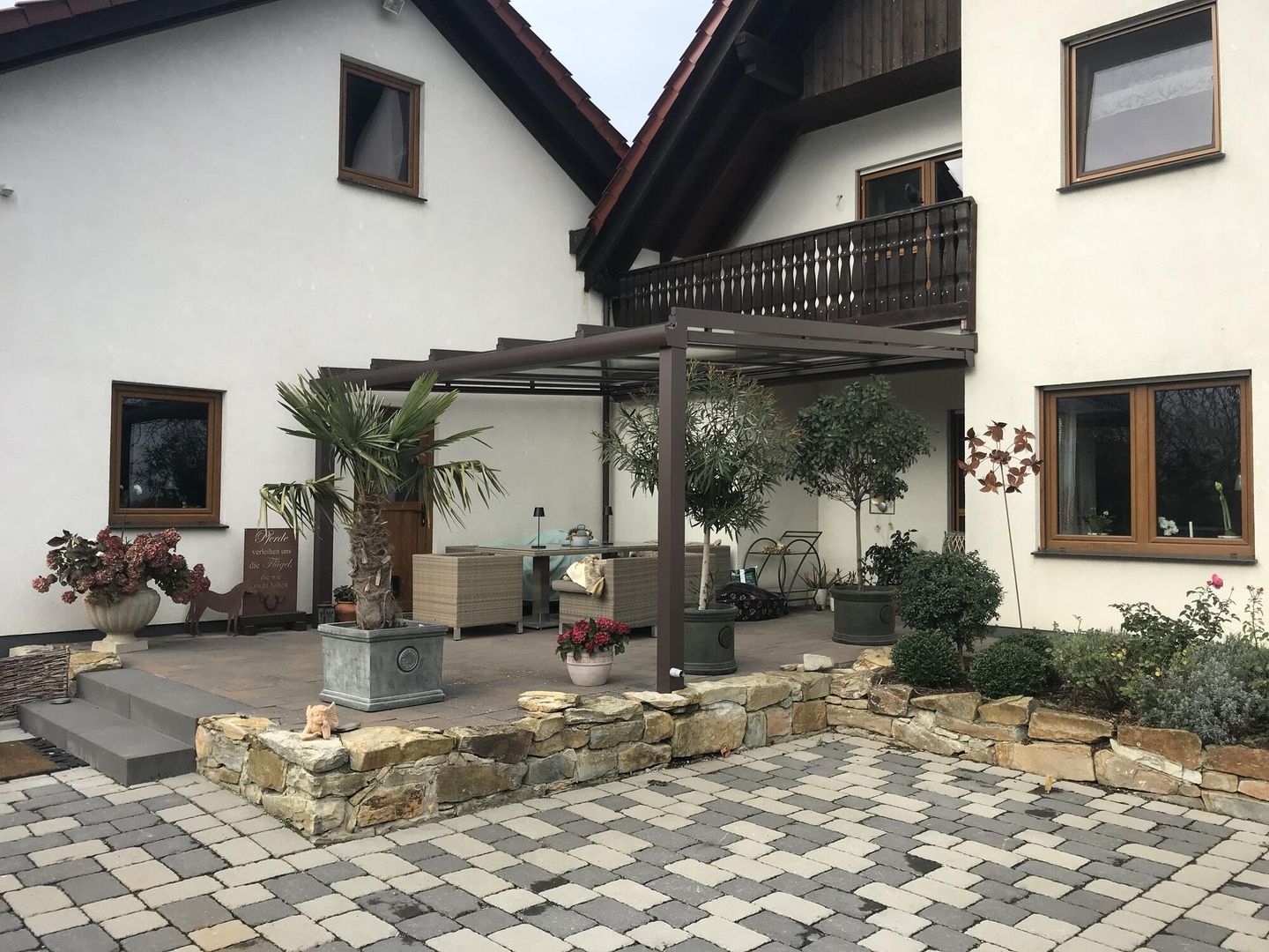 Eine gemütliche Außenterrasse mit Pergola, Grünpflanzen und dekorativen Pflanzen, daneben ein charmantes Haus mit Holzakzenten.