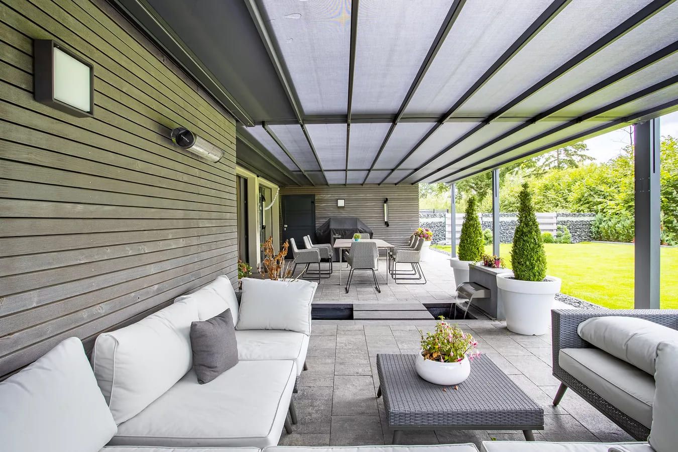 Stilvolle Außenterrasse mit grauen Holzverkleidungen, einer gemütlichen Sitzecke, einem Esstisch, Pflanzgefäßen und einem grünen Rasen unter einem schattigen Dach.