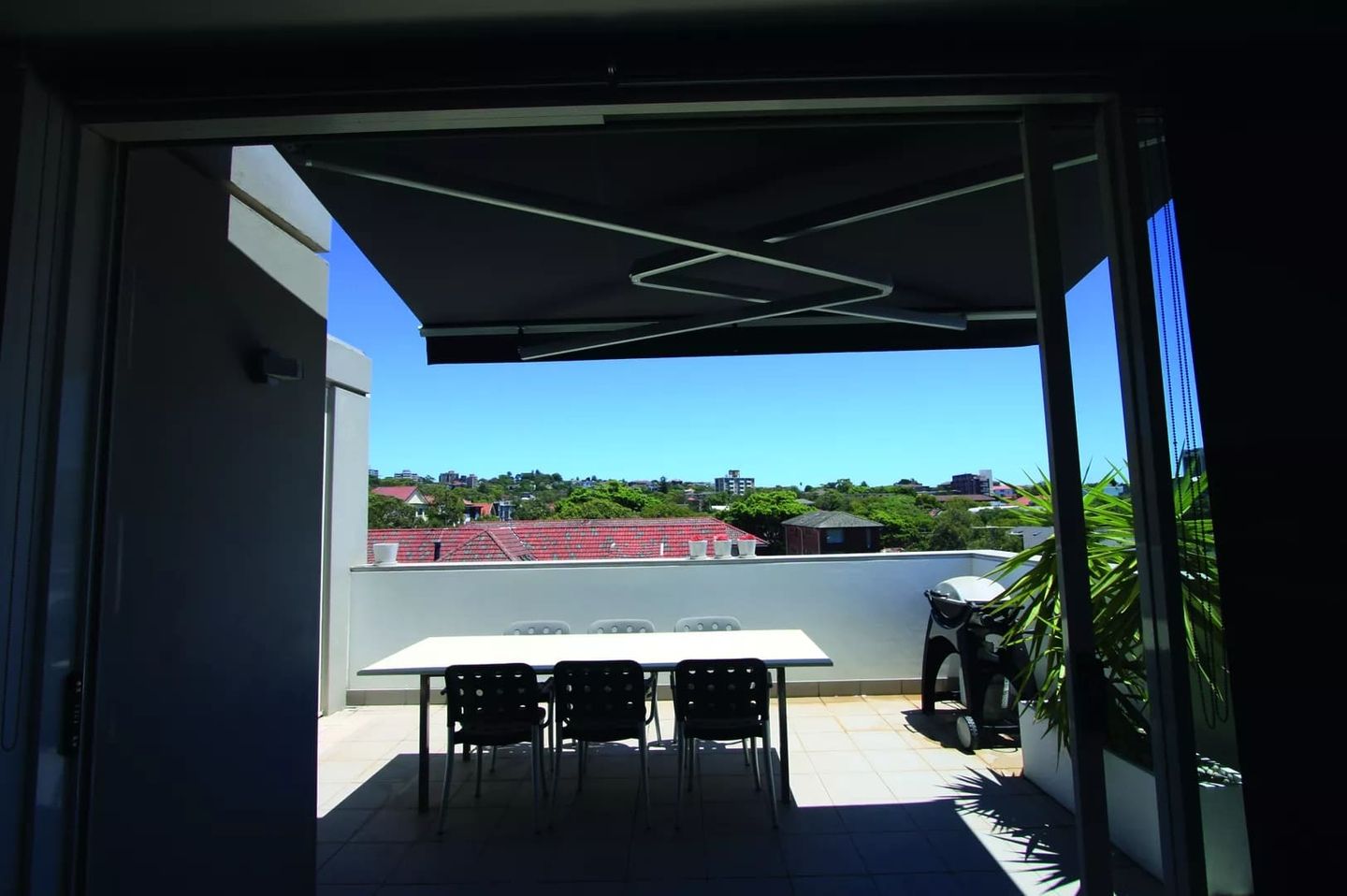 Ein Balkon mit einem rechteckigen Tisch und sechs Stühlen, mit einem Holzkohlegrill, mit Blick auf die Dächer und den blauen Himmel.